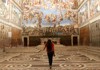 A woman walks through a vast hall in the Vatican Museums, gazing up at the elaborate frescoes covering the ceiling.